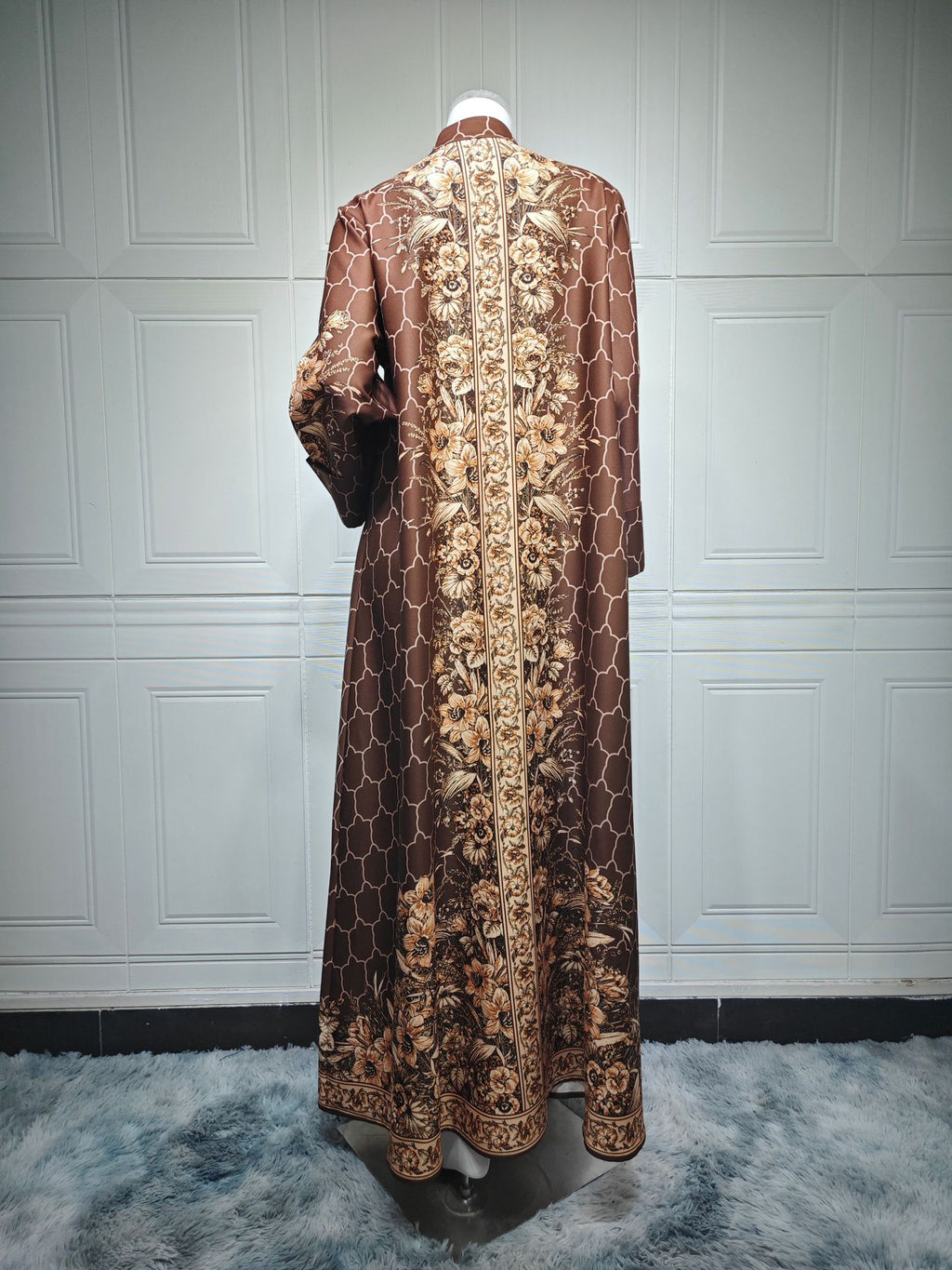 Long, ornate coat with intricate patterns on a mannequin against a white paneled wall.